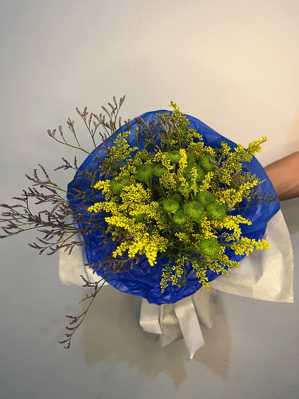 Buquê do Hexa visto de cima com embalagem azul, papel branco e flores em tons de verde, amarelo e lilás